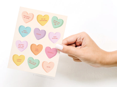 A hand holds the Galentine’s Candy Hearts Greeting Card by Onderkast Studio, featuring twelve colorful candy heart illustrations with fun phrases like BE MY GALENTINE, QUEEN, BAD BITCHES, BESTIES FOR THE RESTIES, and PARTNERS IN WINE.