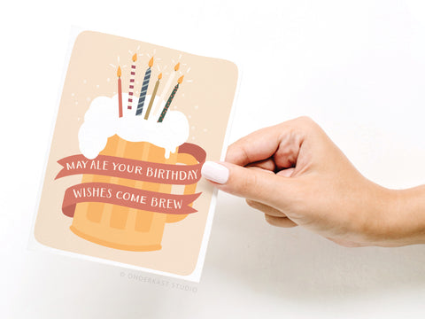 A hand holds Onderkast Studio’s Ale Your Birthday Wishes Greeting Card, featuring a foamy beer mug with five lit candles and a ribbon that says, “May ale your birthday wishes come brew”—perfect for any beer lover.
