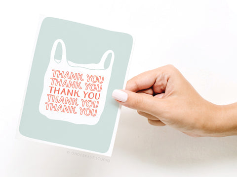 A hand holds Onderkast Studios Thank You Plastic Bag Greeting Card, featuring a white bag with bold red THANK YOU text on a pastel green background—an eco-friendly choice for your greetings.