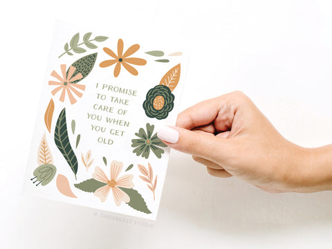 A hand with pale-pink nails holds Onderkast Studios Take Care of You When You Get Old Greeting Card, decorated with green and orange florals. The A2 card reads: I promise to take care of you when you get old.