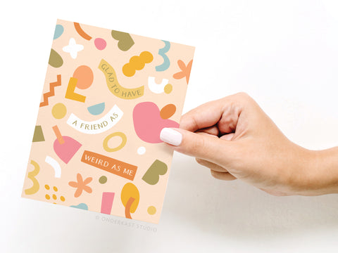 A hand with pale pink nail polish holds the Onderkast Studio Glad to Have a Friend as Weird as Me greeting card, featuring abstract pink, orange, yellow, green, and white shapes on a peach background.