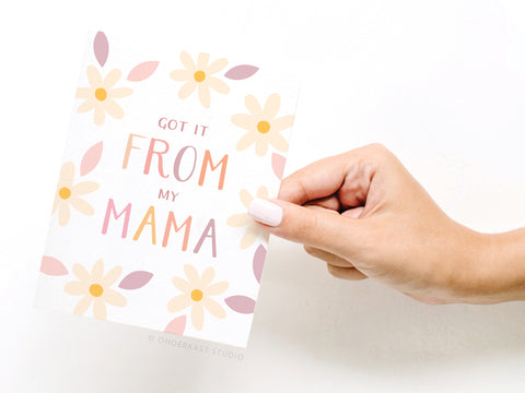 A hand with pale pink nail polish holds the Onderkast Studio Got It From My Mama Greeting Card, featuring pastel flowers and the phrase Got it from my mama in colorful letters.