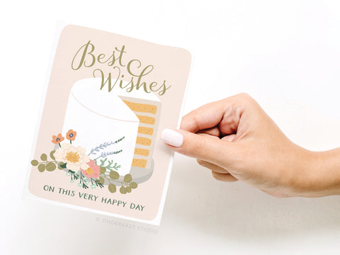 A hand with light pink nail polish holds the Best Wishes Cake Greeting Card by Onderkast Studio, featuring an illustrated cake with flowers and the message: Best Wishes on this very happy day.