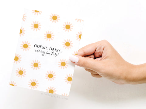 A hand with light pink nail polish holds the Onderkast Studio Oopsie Daisy Belated Birthday Greeting Card, featuring yellow daisies and “OOPSIE DAISY… sorry I’m late!”—a premium choice for belated birthday wishes.