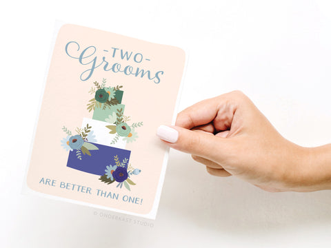 A hand holds the Onderkast Studio Two Grooms Are Better Than One Greeting Card, featuring a decorated three-tier cake, flowers, and a joyful message celebrating love and equality.