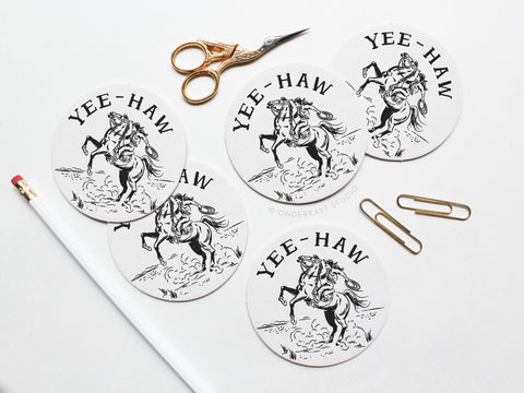 The Yeehaw Cowgirl Coaster Set by Onderkast Studio includes five round, recyclable letterpress coasters featuring a black cowboy on a bucking horse and YEE-HAW text. Gold scissors, paperclips, and a white pencil with red eraser are nearby.