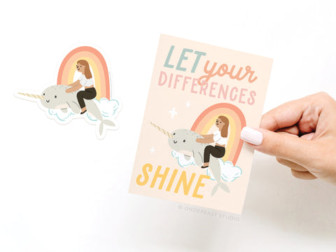 A hand holds Onderkast Studio’s “Let Your Differences Shine Narwhal Sticker Greeting Card,” featuring a girl riding a narwhal on a cloud with a rainbow and the phrase “LET your DIFFERENCES SHINE”—plus a matching vinyl sticker.