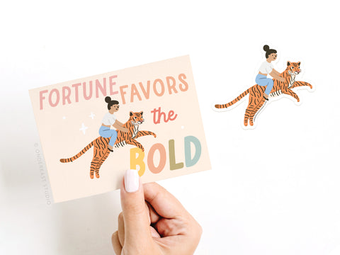 A hand holds Onderkast Studios Fortune Favors the Bold Tiger Sticker Greeting Card, printed on thick white cardstock with a woman riding a tiger. A matching vinyl sticker rests nearby.