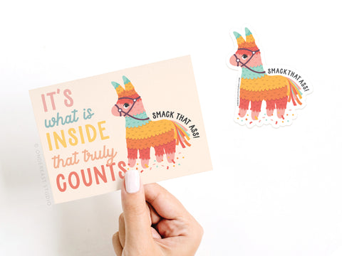 A hand holds Onderkast Studio’s Its What Is Inside That Truly Counts Piñata Sticker Greeting Card, featuring a colorful piñata, the phrase “It’s what is inside that truly counts,” and a matching “SMACK THAT ASS!” vinyl sticker.