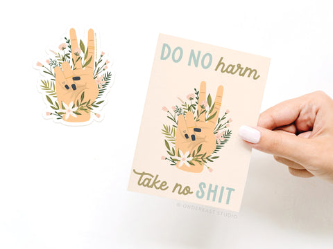 A hand holds the Onderkast Studio Do No Harm Take No Shit Peace Sticker Greeting Card, made of thick white card stock with a floral peace sign and bold text. A matching vinyl sticker is placed next to it on a white surface.