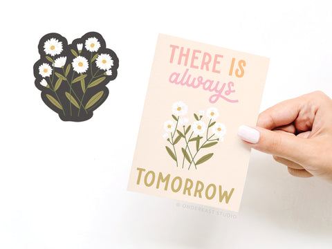 A hand holds the Onderkast Studio There Is Always Tomorrow Flower Sticker Greeting Card, printed on white card stock with illustrated daisies. Next to it lies a matching daisy vinyl sticker on a white background.