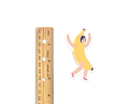 A wooden ruler stands by Onderkast Studios Banana Girl Sticker, a 3-inch tall water-resistant vinyl sticker featuring a person in a banana costume with arms up and pink shoes, shown on a white background.