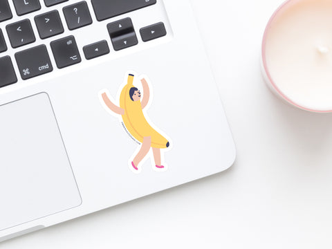 The Banana Girl Sticker by Onderkast Studio, a water-resistant cartoon vinyl, decorates a silver laptop near the touchpad. A pink-rimmed white candle sits nearby on the white surface, highlighting its playful design.