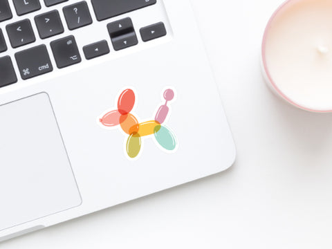 A close-up of a laptop featuring the colorful, water-resistant Balloon Animal Sticker by Onderkast Studio, placed beside a white candle in a pink holder on a white surface.