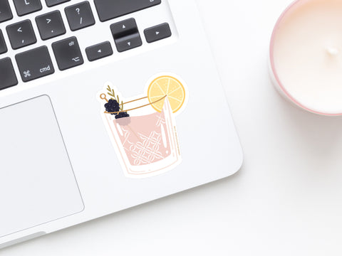 A laptop with the Berry Cocktail Sticker by Onderkast Studio, featuring a decorative drink with a lemon slice and berries, rests on a white surface beside a pale pink candle.