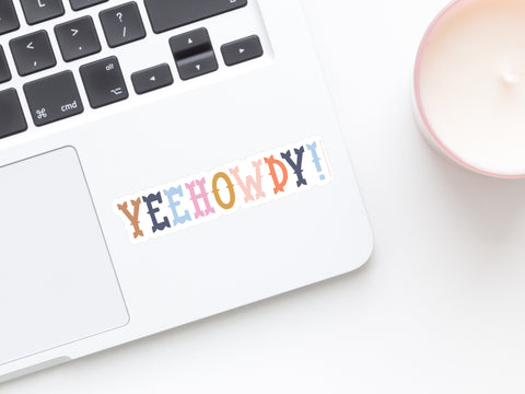 A laptop featuring the Yeehowdy! Sticker by Onderkast Studio, with bold, playful western-style letters and water-resistant design, sits beside a pink candle on a white surface.