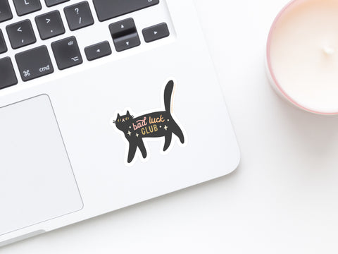 A laptop features the Onderkast Studio Bad Luck Club Sticker—a weatherproof black cat decal—on its keyboard area, while a lit candle in a pink holder rests nearby on a white surface.