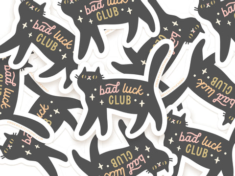 Multiple Bad Luck Club Stickers by Onderkast Studio, featuring playful black cats and stars, are scattered on a white background. Each weatherproof sticker has “bad luck CLUB” in pink and yellow cursive text.