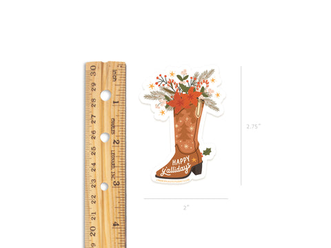 The Happy Yallidays Cowboy Boot Sticker by Onderkast Studio features a festive boot with greenery and flowers, measuring about 2.75 inches tall and 2 inches wide, as shown next to a wooden ruler. Water-resistant and decorative!.