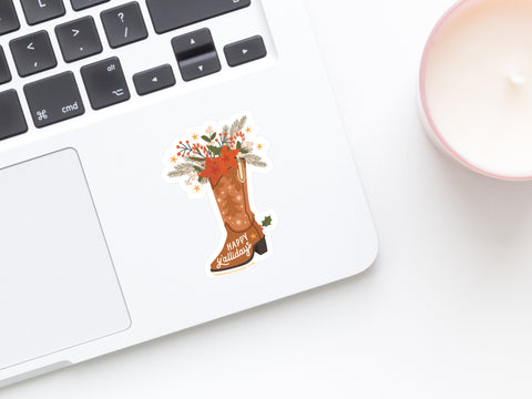 A laptop with Onderkast Studio’s Happy Y’allidays Cowboy Boot Sticker—a durable vinyl decal featuring a brown boot filled with flowers, leaves, and Happy Holidays—sits beside a white candle in a pink holder on a white surface.