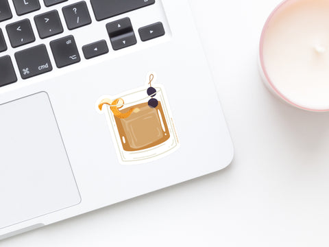 A laptop features the Onderkast Studio Old Fashioned Cocktail Sticker, showing a glass with a brown drink, orange slice, and cherries on a cocktail stick. Nearby is a white candle in a pink holder, all placed on a white surface.