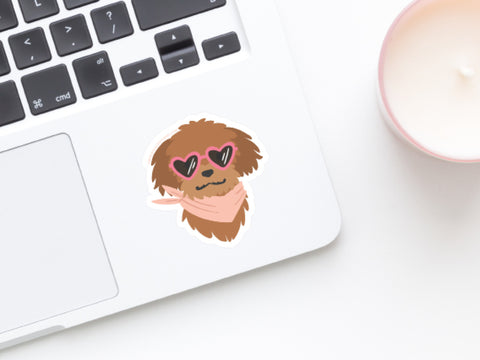 A laptop with the Onderkast Studio Dog w/ Heart Sunglasses Sticker on the palm rest, next to a white candle in a pink container.