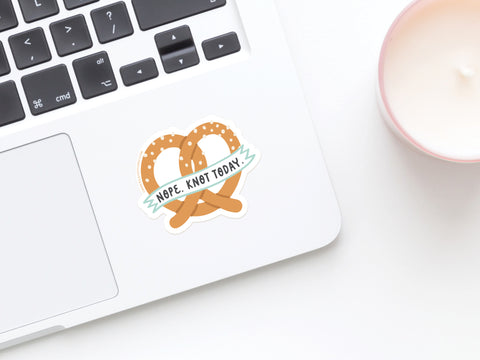 A laptop features the Onderkast Studio Nope Knot Today Pretzel Sticker. A lit white candle in a pink container is nearby on a white surface.