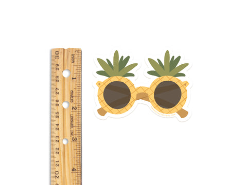 A wooden ruler beside the Pineapple Sunglasses Sticker by Onderkast Studio—a water resistant, durable vinyl sticker featuring yellow pineapple-textured frames and green tops. Measures about two inches wide.