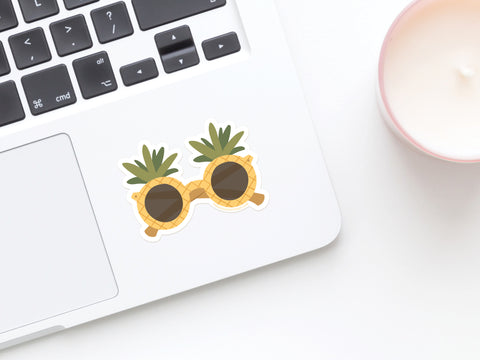 A close-up of a laptop keyboard with the Onderkast Studio Pineapple Sunglasses Sticker—a durable, water-resistant vinyl decal—on the palm rest, beside a white candle in a pink holder on a white surface.