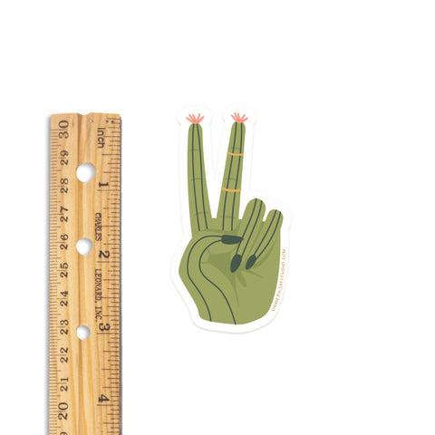 The Cactus Peace Sign Sticker by Onderkast Studio is a durable, water-resistant vinyl sticker featuring a green hand with black nails and cactus-like fingertips, measuring about 3 inches tall.
