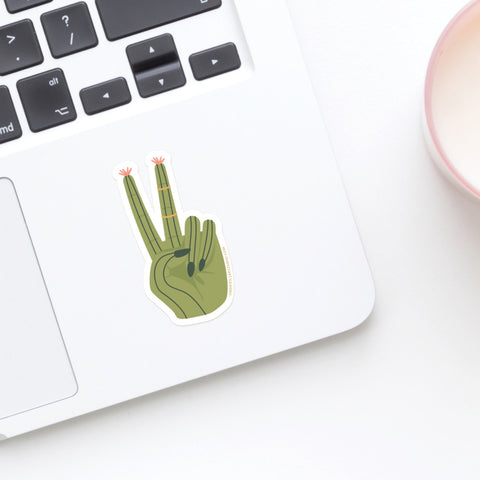 A laptop is decorated with the Onderkast Studio Cactus Peace Sign Sticker near the keyboard, while a cup holding a light drink sits partially in view on the right.