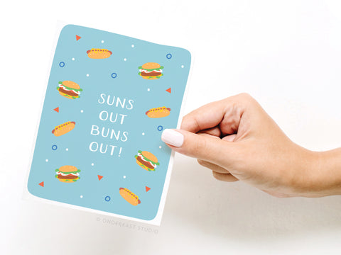 A hand with light skin holds the Onderkast Studio Suns Out Buns Out! Greeting Card – FINAL STOCK, featuring burgers, hot dogs, and bold text on a blue background with small geometric shapes.