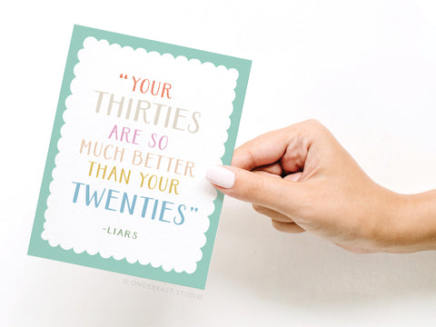 A hand holds Onderkast Studios Your Thirties Greeting Card, featuring scalloped white center, teal border, and colorful text: Your thirties are so much better than your twenties – Liars.