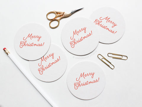 The Merry Christmas Coaster Set by Onderkast Studio features round white letterpress coasters with red text, arranged with gold scissors, a white pencil, and paperclips—ideal for gifting in a recyclable kraft box.