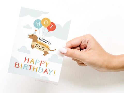 A hand holds the Hot Diggity Dog Greeting Card by Onderkast Studio, featuring a cartoon dachshund with HOT balloons above DIGGITY DOG and HAPPY BIRTHDAY! in colorful letters on a cloudy sky background.