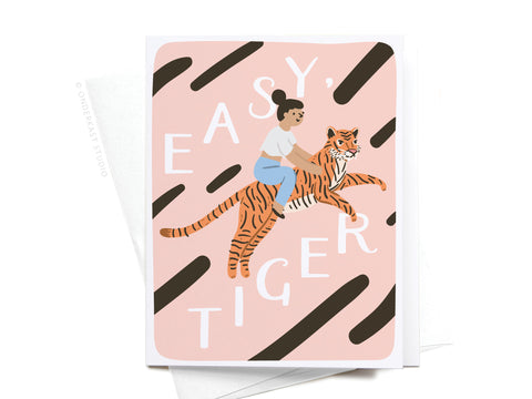 Featuring an illustration of a woman riding a tiger on pink with dark stripes, the Easy, Tiger Greeting Card – FINAL STOCK by Onderkast Studio brings playful style and premium quality to any card collection. White text reads “EASY, TIGER” in a fun font.