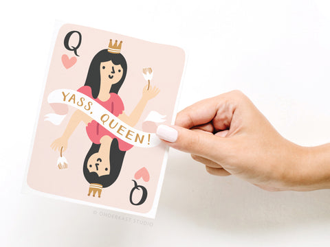 A hand with pale pink nails holds the Onderkast Studio Yass, Queen! Greeting Card – FINAL STOCK. This empowering friendship card features a crowned Queen of Hearts with cake, a gold YASS, QUEEN! banner, and a light pink background.