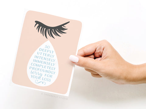 A hand with pale pink nail polish holds the So Sorry for Your Loss Teardrop Greeting Card by Onderkast Studio, a heartfelt condolence card featuring a large eyelash above a teardrop and the message: So deeply, utterly, intensely sorry for your loss.