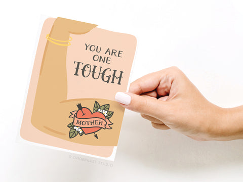 A hand holds the You Are One Tough Mother Greeting Card by Onderkast Studio, featuring an illustrated flexed arm with a heart tattoo that says Mother, plus vibrant colors and bold lettering for a premium look.