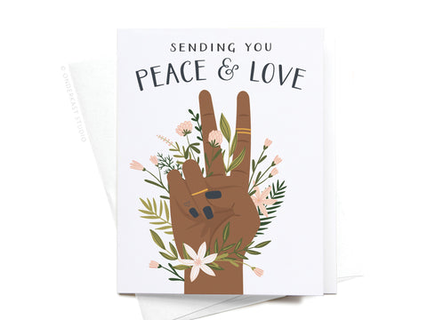 The Sending You Peace & Love Greeting Card by Onderkast Studio features a dark hand making a peace sign with black nails, gold rings, and pastel flowers, plus the message “Sending You Peace & Love” above. Color: Dark.