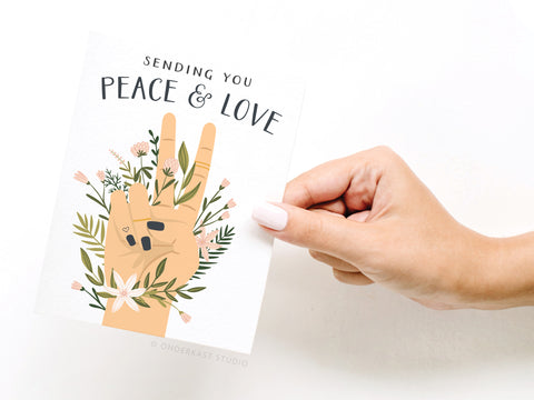 A hand with light pink polish holds Onderkast Studio’s Sending You Peace & Love Greeting Card, featuring an illustrated peace sign with flowers. The card reads: “Sending you peace & love”—a premium thinking of you card.