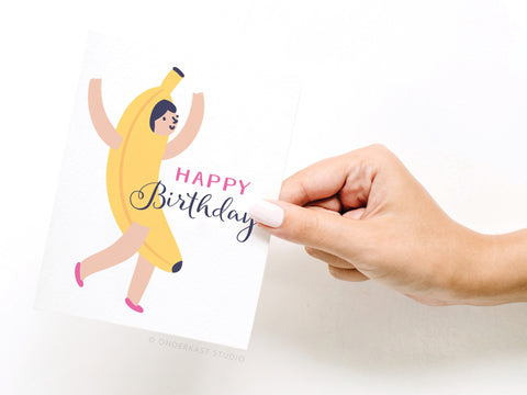 A hand holds the Happy Birthday! Banana Suit Girl Greeting Card by Onderkast Studio, featuring an illustration of Banana Suit Girl with arms raised and Happy Birthday in pink and black letters.