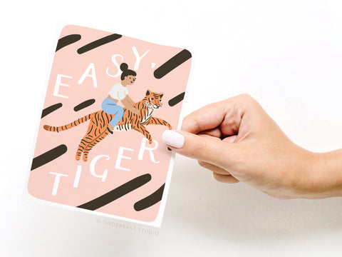 A hand with white nail polish holds the Easy, Tiger Greeting Card – FINAL STOCK by Onderkast Studio, featuring a woman riding a tiger with Easy, Easy, Tiger on a pink background—an everyday card with premium feel.