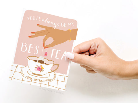 A hand with light pink nail polish holds the Onderkast Studio You’ll Always Be My Bestea Greeting Card, featuring a teacup illustration—an ideal card for any tea-loving friend.