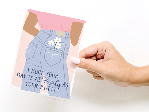 A hand holds the Onderkast Studio I Hope Your Day Is As Lovely As Your Butt greeting card, featuring a cartoon in jeans with daisies. Printed in the USA, this playful card gives a cheeky compliment.