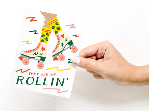A hand with white nail polish holds the Onderkast Studio They See Me Rollin Roller Blades Greeting Card, featuring legs in yellow-patterned socks and pink skates, colorful doodles, and the phrase THEY SEE ME ROLLIN’—perfect for skating fans.