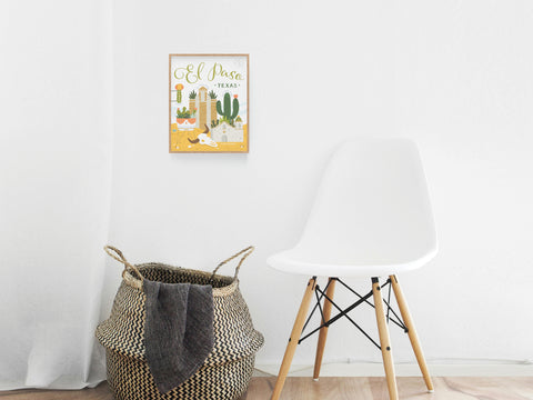 A white chair and woven basket sit against a white wall, topped by the El Paso Succulent Skyline Art Print from Onderkast Studio, featuring cacti, a guitar, hat, and desert-inspired decor.