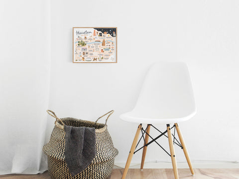 A woven basket with a folded blanket sits next to a modern white chair on wooden flooring, under the Houston Map Art Print by Onderkast Studio, which hangs framed on a white wall and features illustrated neighborhoods.