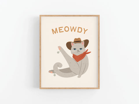 An illustration of a gray cat in a cowboy hat and red bandana, raising one leg under MEOWDY. The framed Meowdy Cat Art Print by Onderkast Studio is displayed on a white wall—ideal for cat lovers and art print fans.
