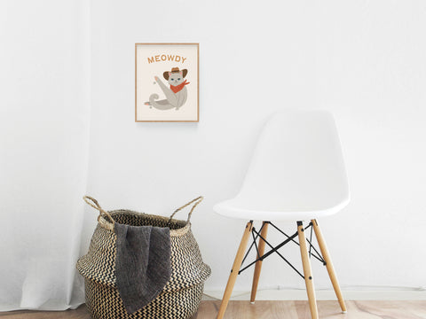 A white modern chair sits beside a woven basket on a wooden floor, while the framed Meowdy Cat Art Print by Onderkast Studio adds whimsical feline charm to the white wall—ideal for cat lovers and art enthusiasts.